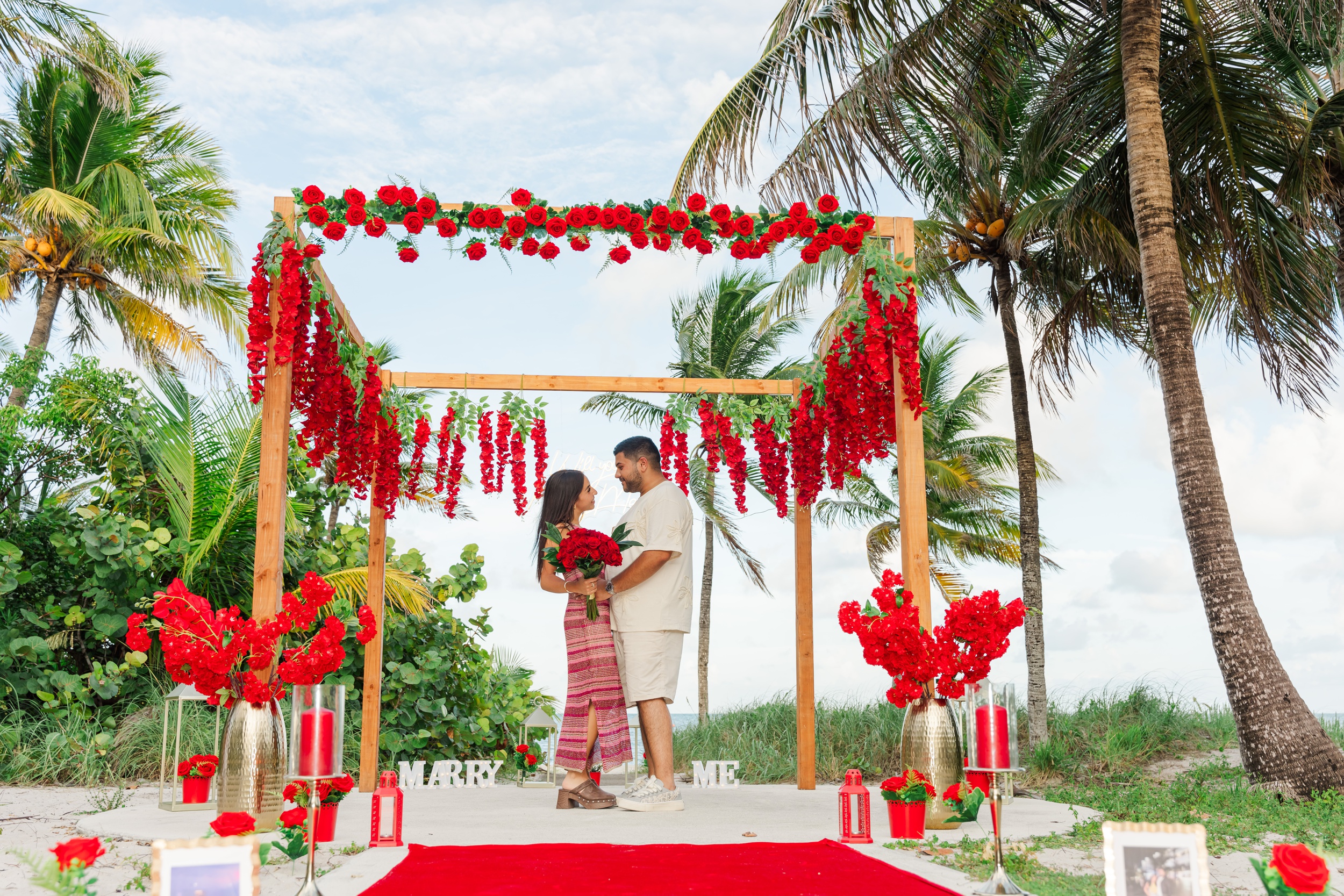 A happy newly engaged couple stands in the center of an elaborate red floral setup on a beach