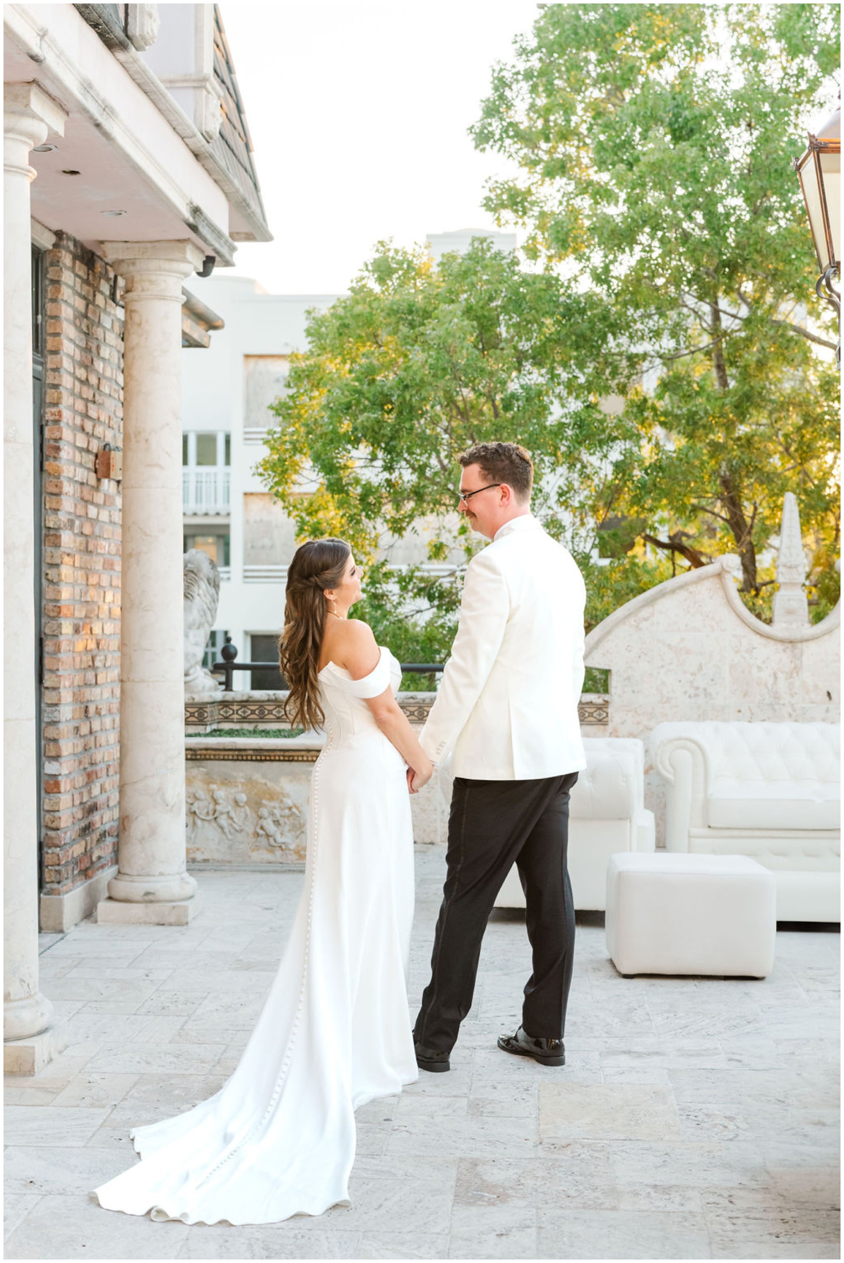 Newlyweds hold hands while hanging out on a patio in a white suit jacket and long white gown