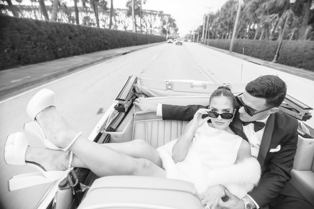 A bride kicks back with feet out the side of a convertible leaning on her groom while riding in the back seat