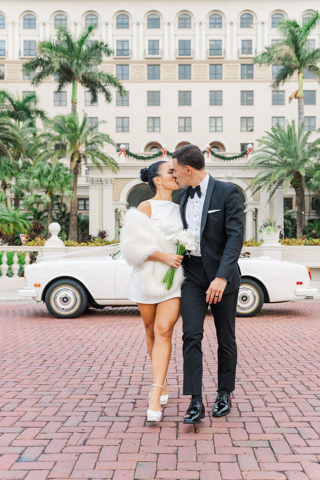 Newlyweds walk and kiss while walking on a brick driveway in front of a resort