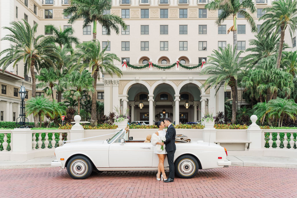 Newlyweds kiss while leaning against a vintage white car in front of one of the wedding venues in west Palm Beach