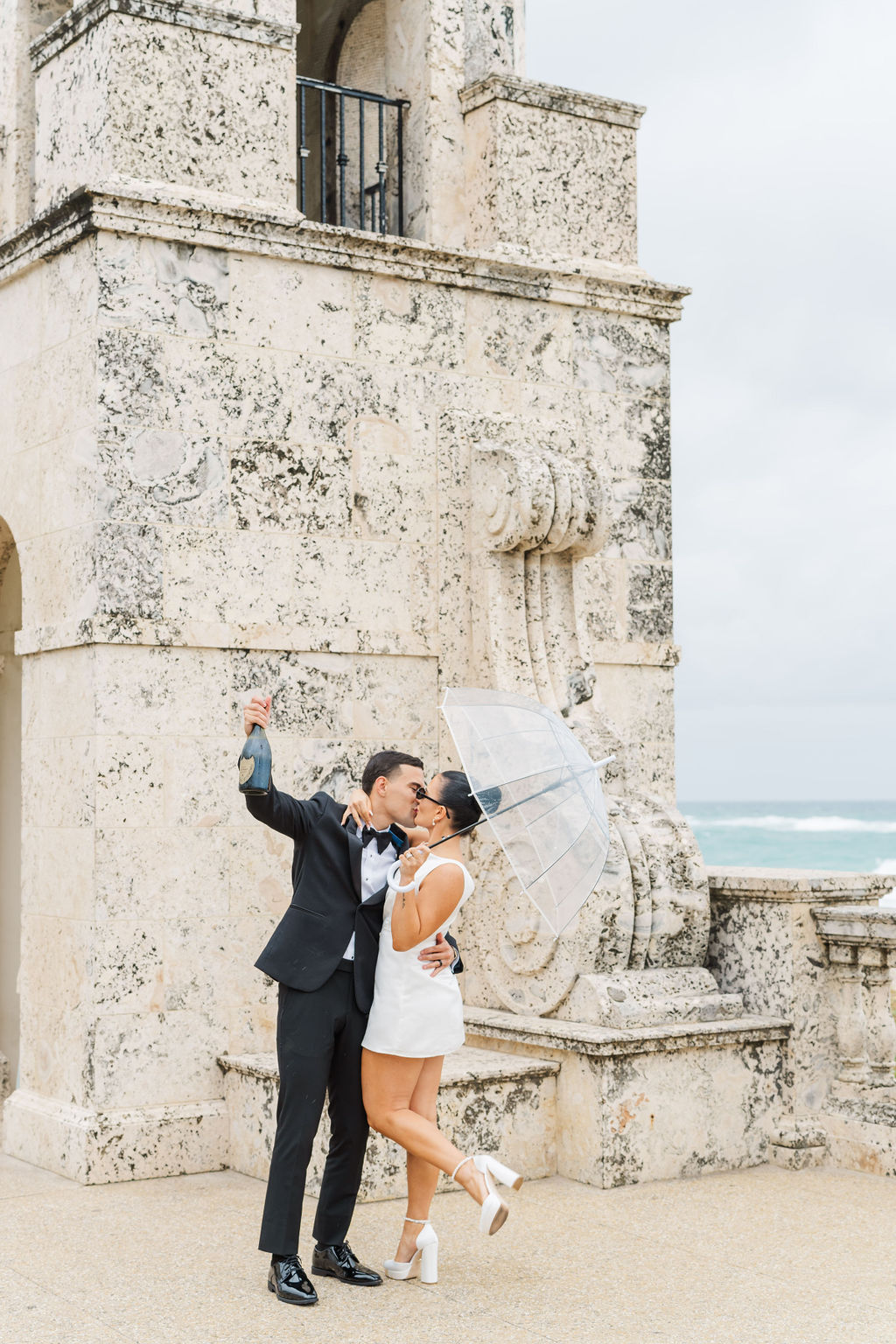 Newlyweds kiss with a clear umbrella and bottle of champagne at one of the wedding venues in west Palm Beach