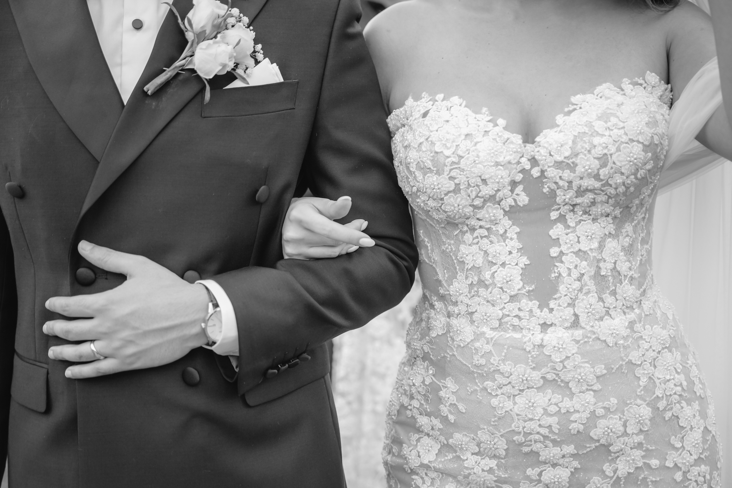 Details of a bride's lace dress and groom's black suit and boutonniere as they lock arms