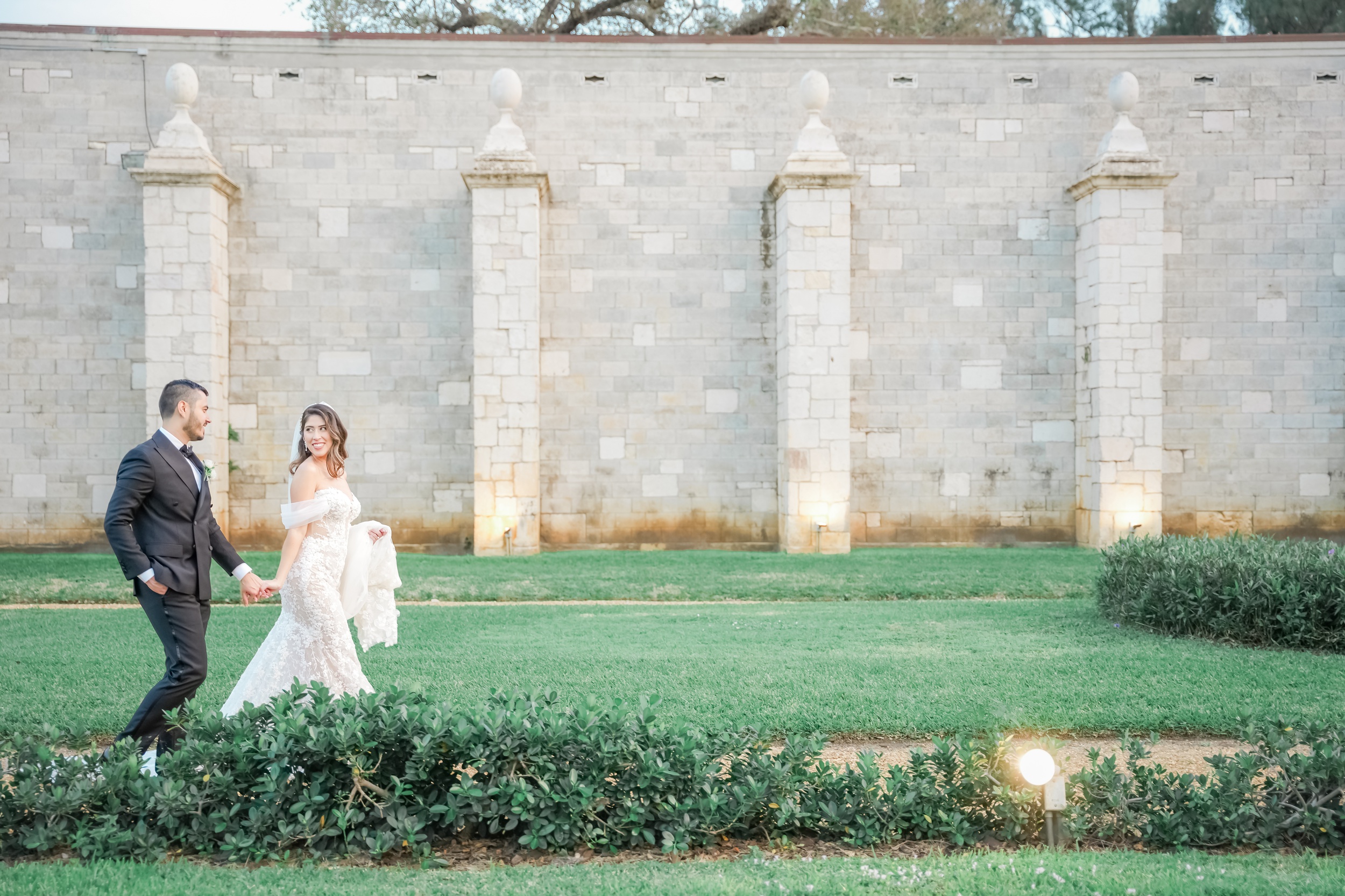 A bride leads her groom by the hand through a garden path and smiles back at him