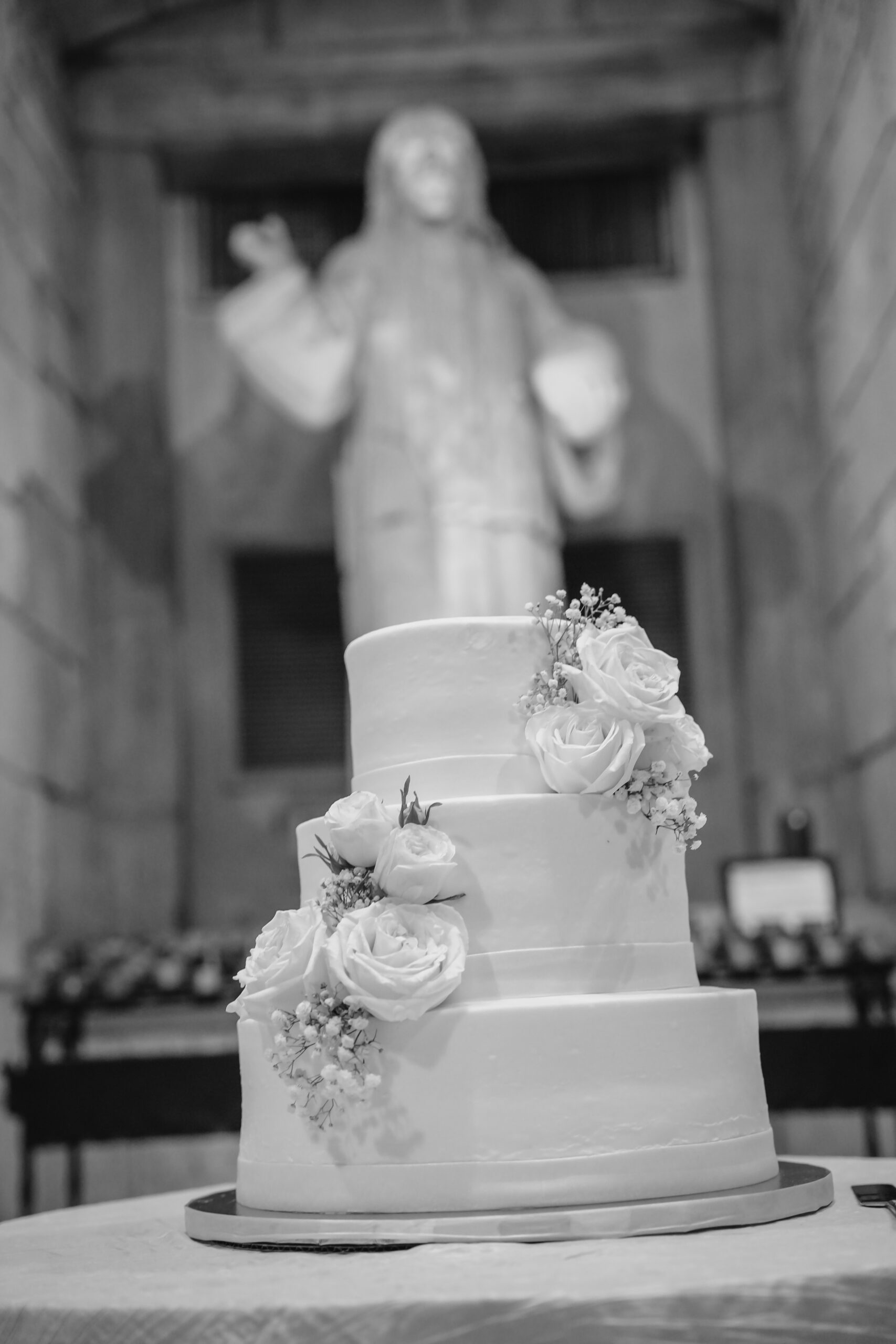 A three tier cake sits on a table under a large statue at the Ancient Spanish monastery wedding venue