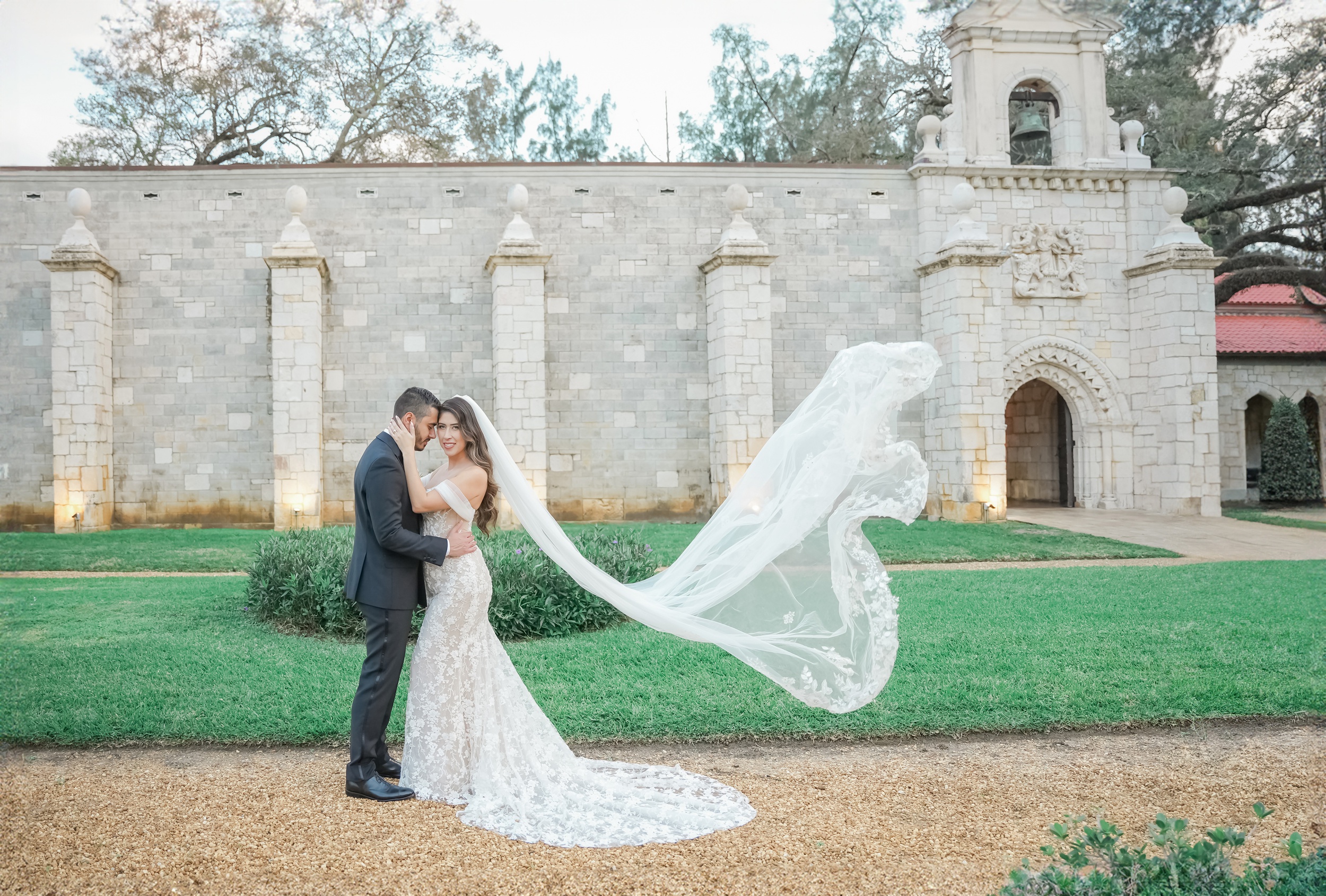 A bride's veil flies behind her in the wind as she snuggles her groom in a garden at one of the european style wedding venues in florida