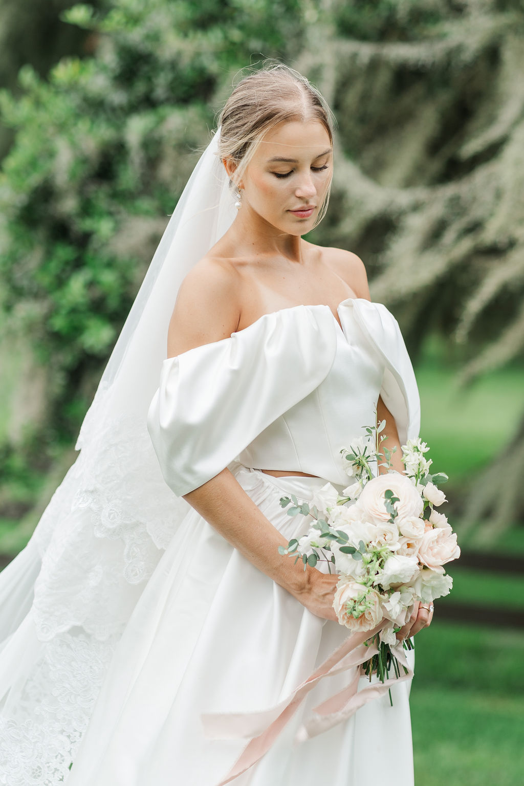 A bride walks in a garden looking down at her white bouquet in her hands