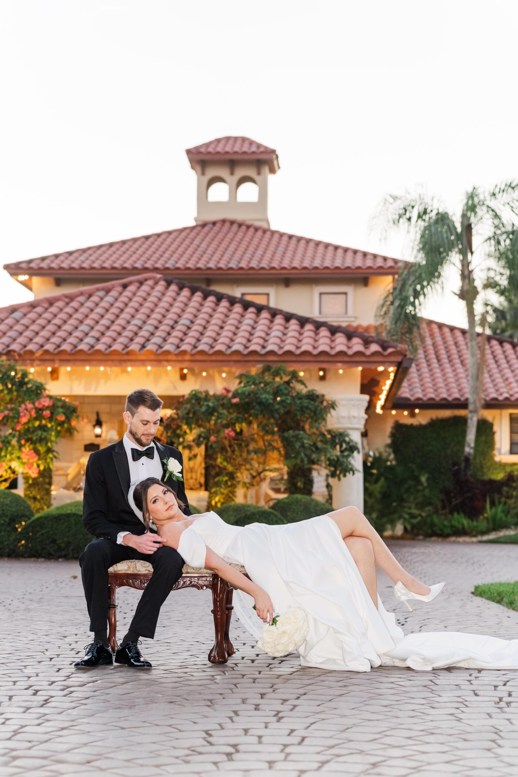 A bride lays in the lap of her groom in the driveway on a vintage bench at one of the luxury wedding venues in miami